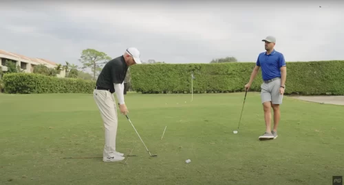 Two Performance Golf coaches on the green, one on the left is in the golf short game setup while the other is on the right, leaning on his golf club watching on.