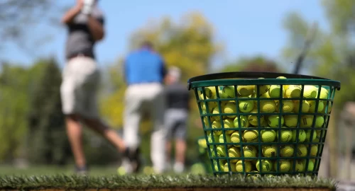 Close-up of a bucket full of yellow golf balls, with various golfers in the background practicing their golf swings.