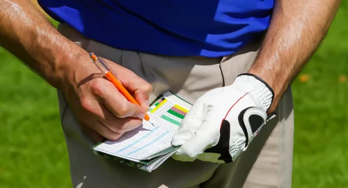 A close up shot of a man wearing one golf glove on his left hand, with his ungloved right hand writing a golf score on a golf scorecard.