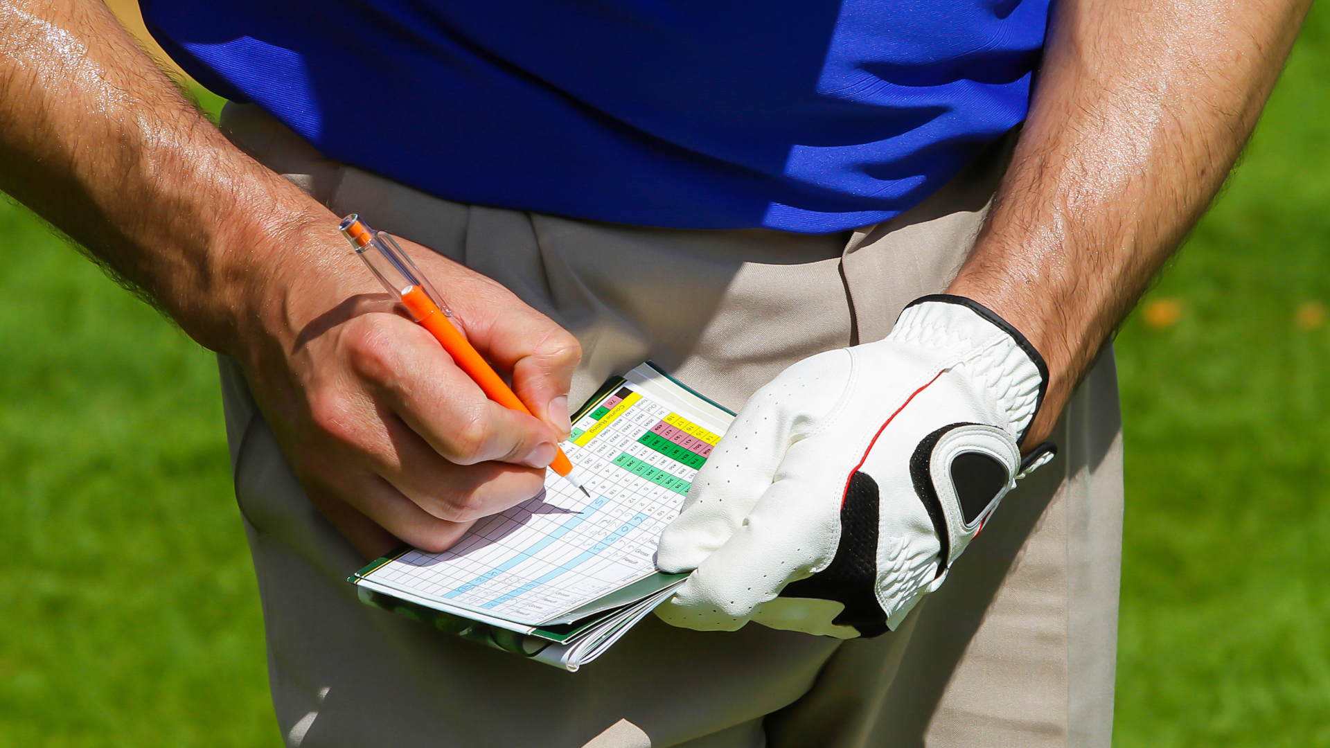 A close up shot of a man wearing one golf glove on his left hand, with his ungloved right hand writing a golf score on a golf scorecard.